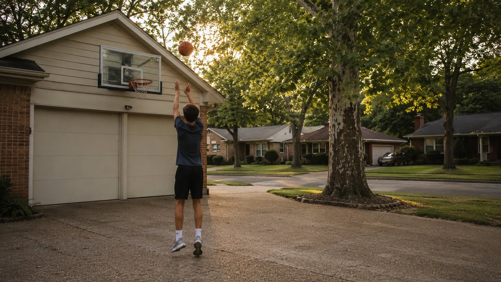 A teenager shooting basketball in a driveway in a mid-century Memphis neighborhood in late afternoon light, with neighboring homes and a mature sycamore tree visible.